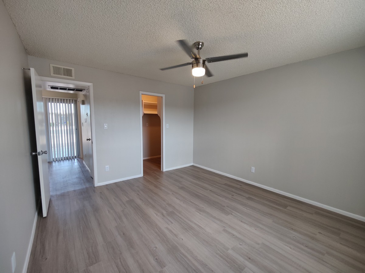 An empty room with wood flooring and a ceiling fan, featuring a doorway leading to another area.