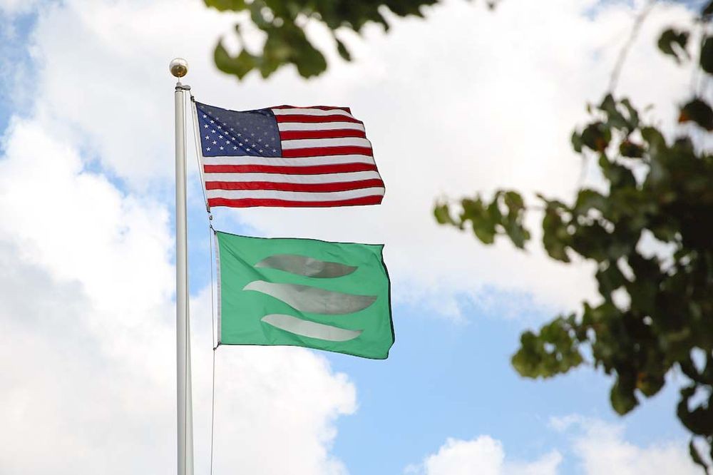 An American flag and a green flag flying on a flagpole against a backdrop of sky and clouds.