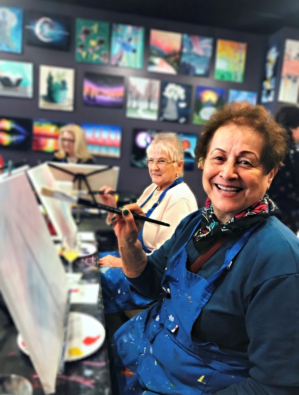 A group of people enjoy painting together in an art class, with one person smiling at the camera.