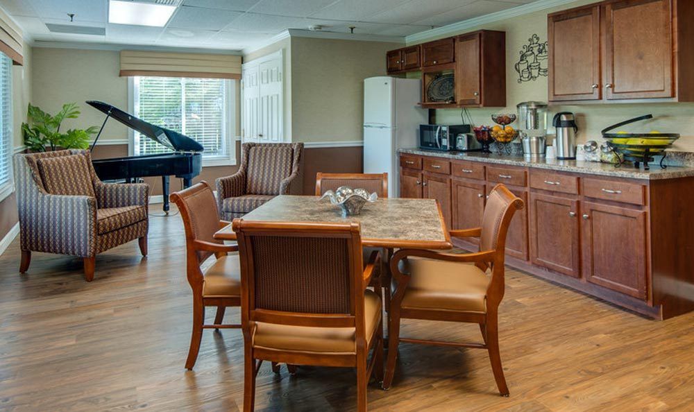 A cozy dining area with a kitchen in the background, featuring a table with chairs and a piano by the window.