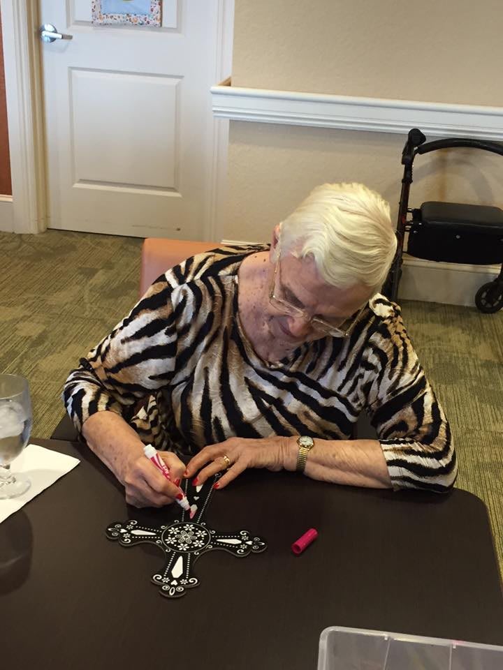 An elderly woman is sitting at a table, creating artwork with a black marker on a black and white patterned paper.