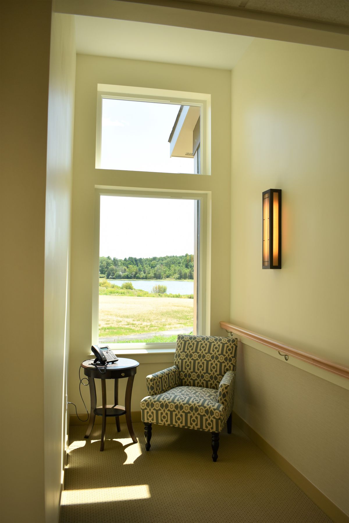 A cozy reading nook by a large window with natural light streaming in, featuring a patterned chair, side table, and wall lamp.