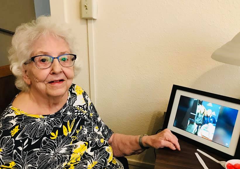 An elderly woman sitting by a desk, smiling and pointing to a photo in a frame.