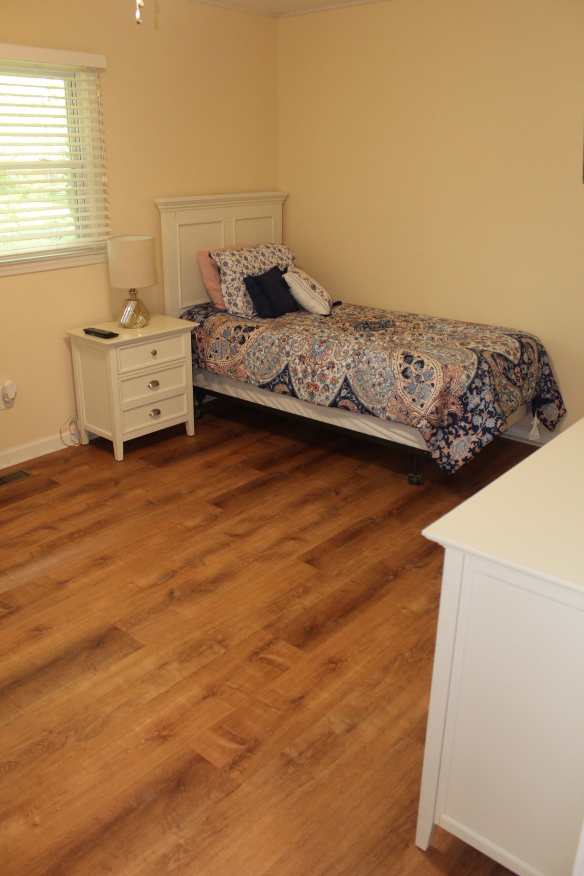 A simple bedroom with a single bed, nightstand, and dresser on wooden flooring.