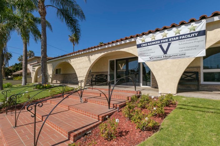 A sunny day at the entrance of a Mediterranean-style building with palm trees and well-maintained landscaping.