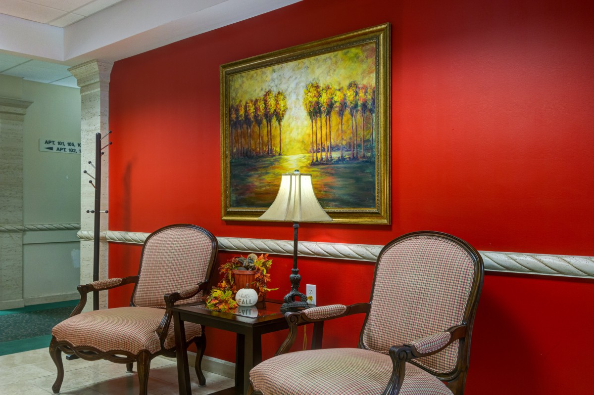 A cozy sitting area with two chairs, a table, and a lamp against a vibrant red wall adorned with a colorful painting.
