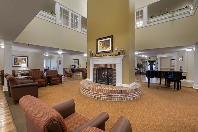 A cozy living room with a central fireplace, comfortable seating, and a grand piano in the background.