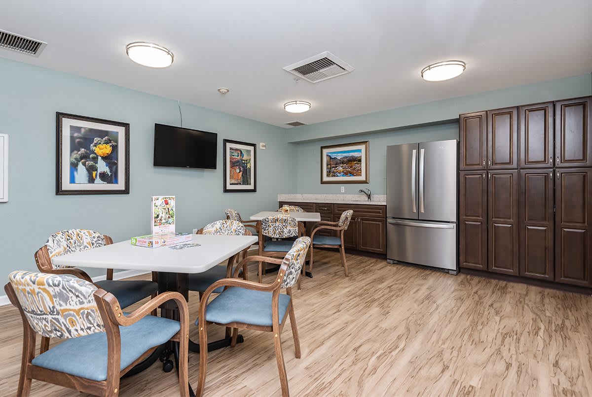 A cozy dining area with tables, chairs, and a refrigerator in the corner, adorned with artwork on light blue walls.