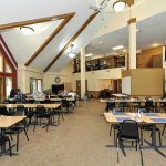 A spacious classroom with tables and chairs arranged under a high ceiling.