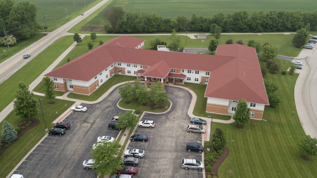 Aerial view of a large building with a red roof and surrounding parking lot, set in a landscaped area near a road.