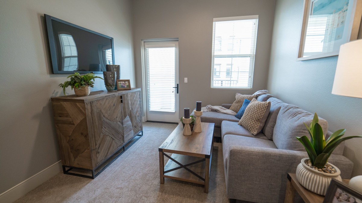 A cozy living room with a gray sofa, wooden TV stand, and a coffee table in front of a window and door.