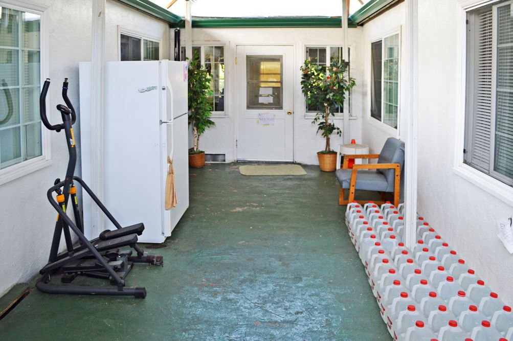A covered patio with a refrigerator, seating area, and exercise equipment, lined with water bottles and potted plants.