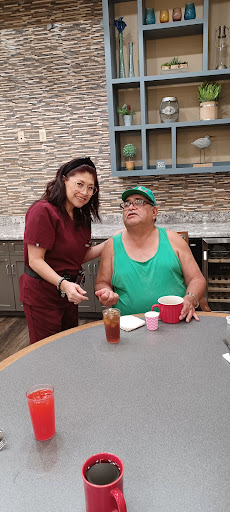 A woman in scrubs serves a drink to a man in a green tank top at an outdoor dining setting.