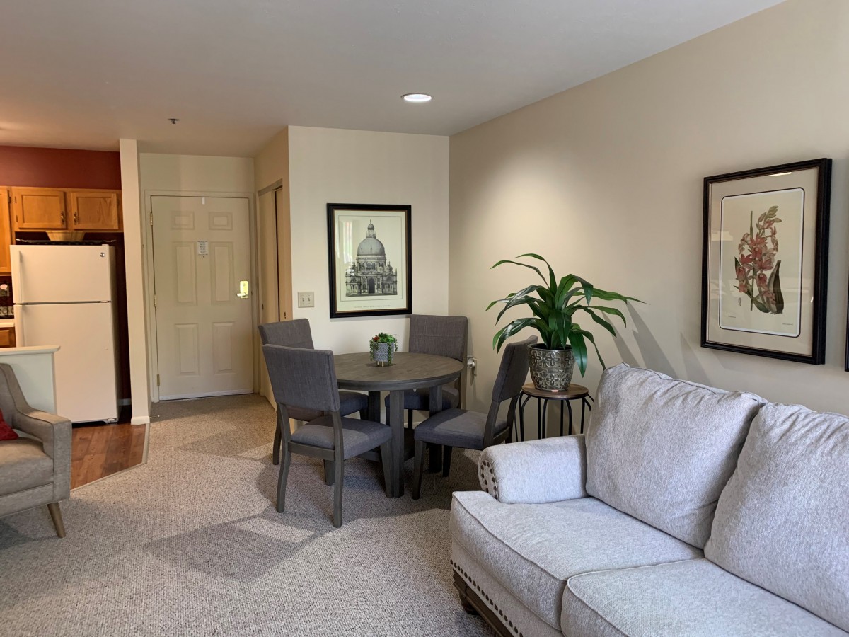 A cozy living room with a small dining area, featuring a sofa, chairs, a round table, indoor plant, and framed artwork on the walls.