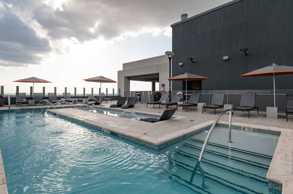 A modern rooftop pool with sun loungers and umbrellas overlooks an urban skyline under a cloudy sky.