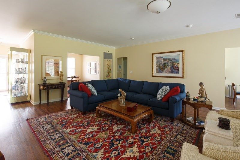 A cozy living room featuring a blue sectional sofa, a vibrant patterned carpet, and a wooden coffee table. Walls are adorned with framed artwork and the space is warmly lit.
