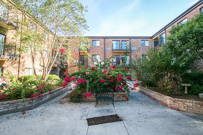 A scenic courtyard featuring a brick apartment building, lush greenery, and vibrant flowers with a central bench.