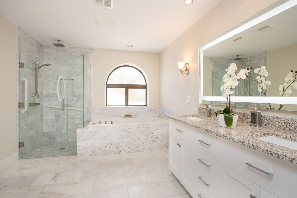 A modern, luxurious bathroom featuring a marble bathtub and glass shower with a vanity and illuminated mirror.