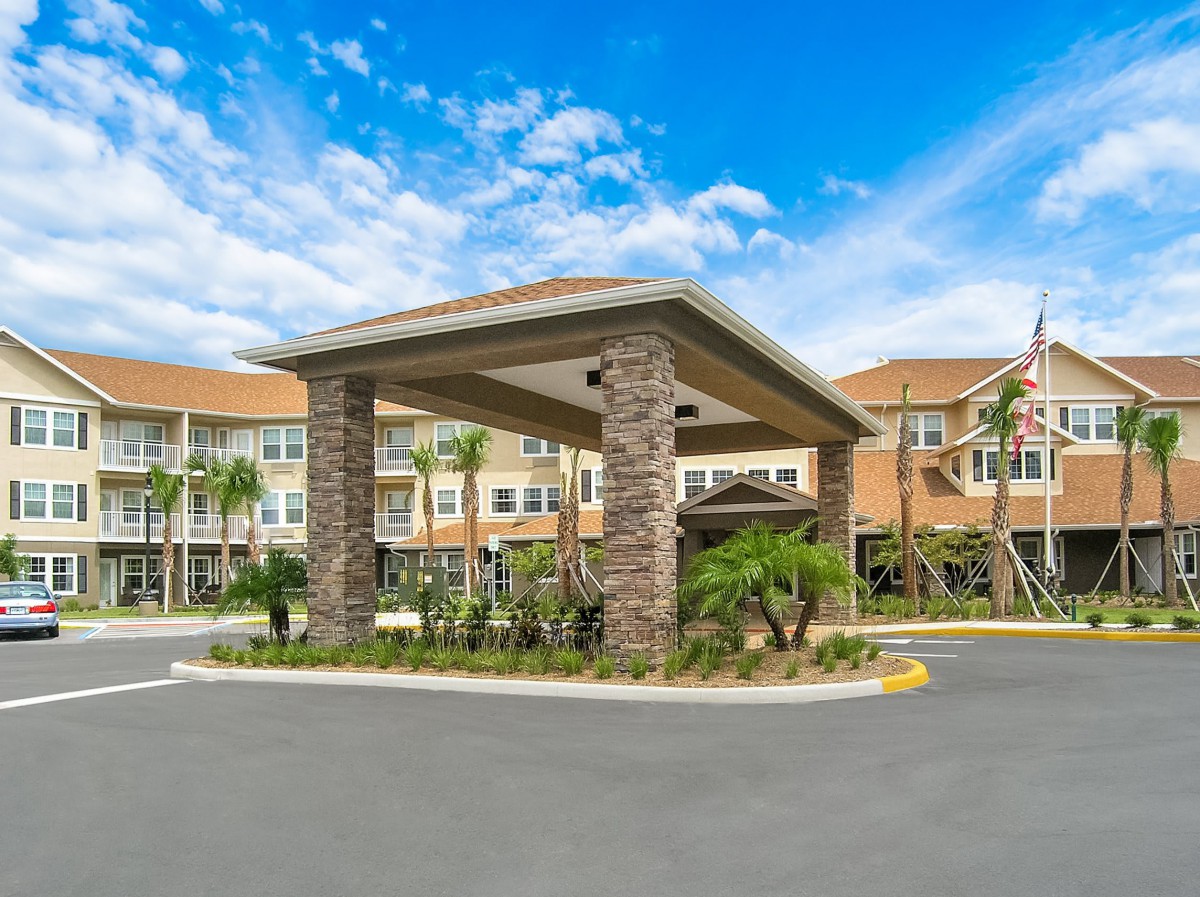 A modern building entrance with a covered driveway and surrounding palm trees under a partly cloudy sky.