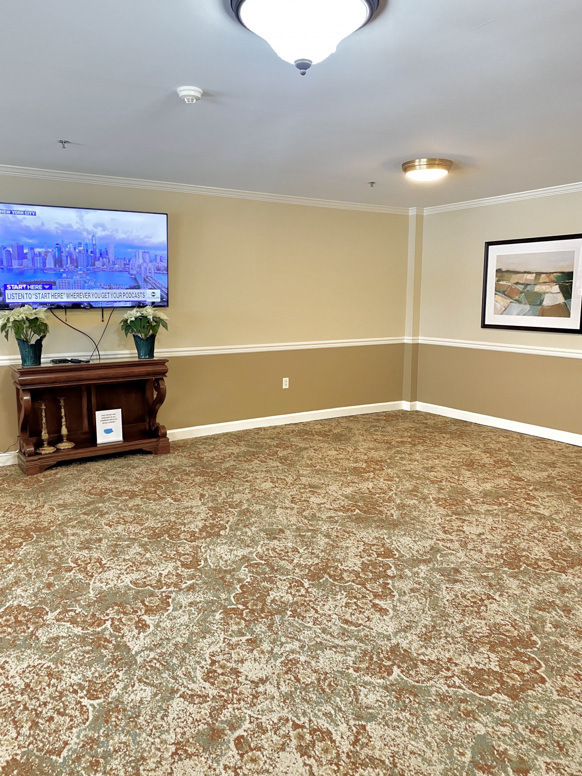 An empty living room with a carpeted floor, featuring a wall-mounted television, a painting, and some furniture.