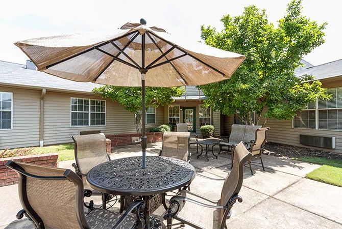 A sunny patio with outdoor furniture including a table, chairs, and an umbrella surrounded by trees.