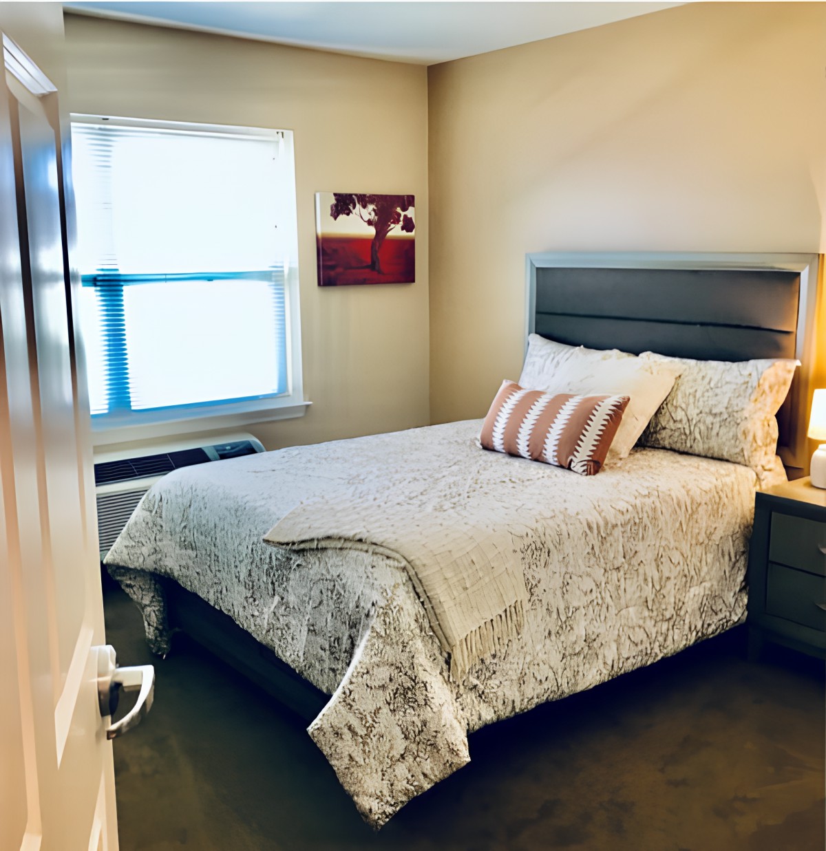 A cozy bedroom with a neatly made bed, featuring patterned bedding and decorative pillows. The room is softly lit by a bedside lamp, with a window allowing natural light to brighten the space.