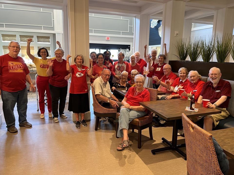 A group of seniors gathered together, some sitting and others standing, all wearing red clothing in a restaurant setting.