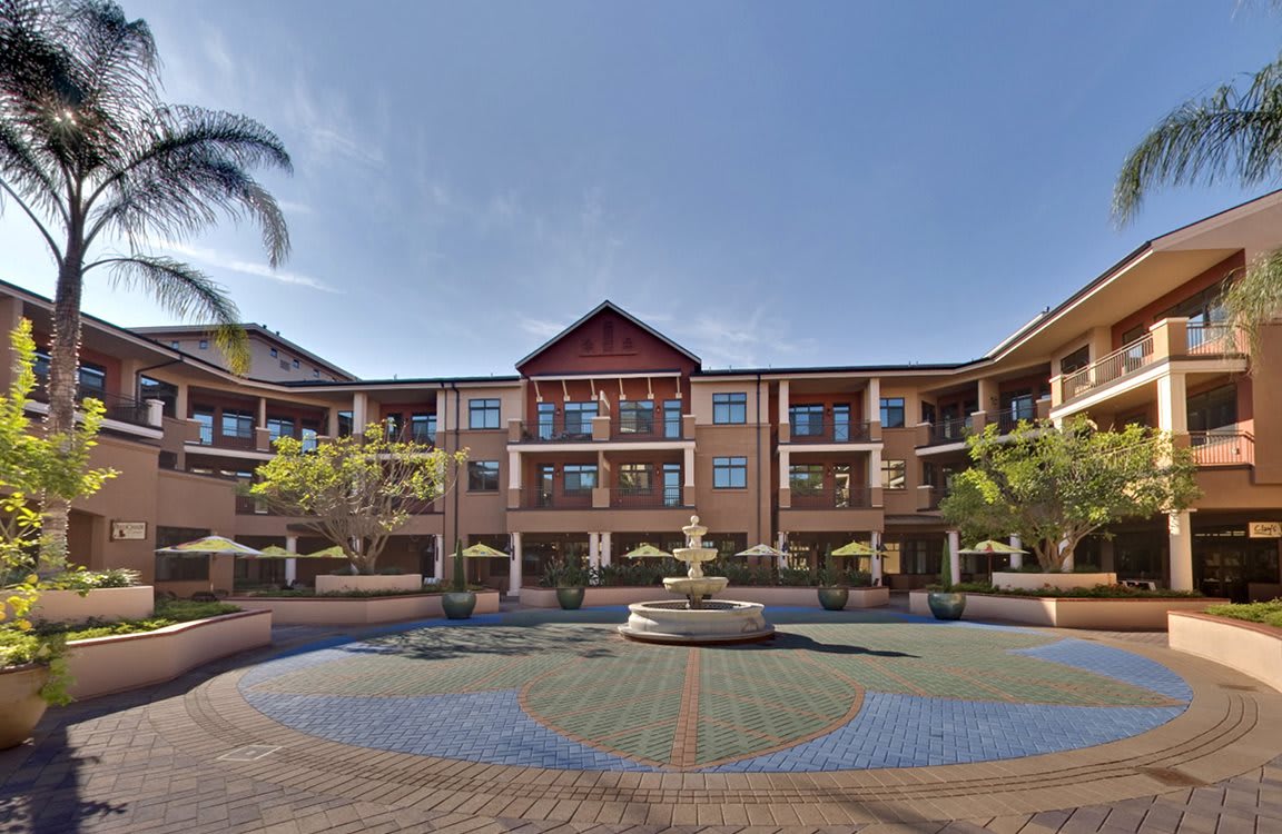 A sunny courtyard featuring a brick building with balconies and a central fountain, surrounded by trees.
