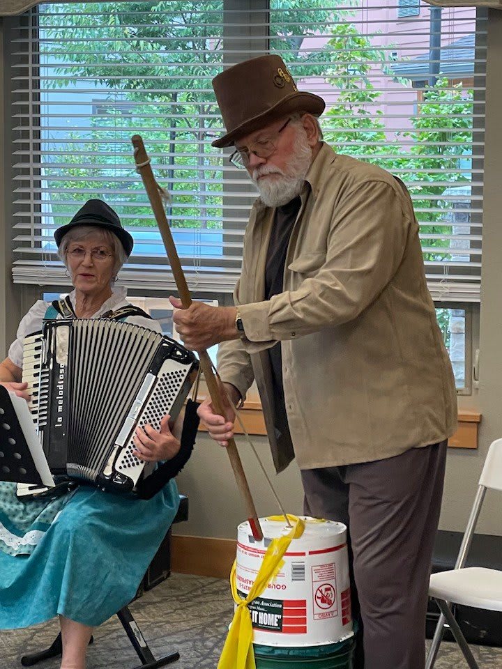 Two musicians perform indoors; one plays the accordion while the other uses a makeshift instrument with a bucket and stick.