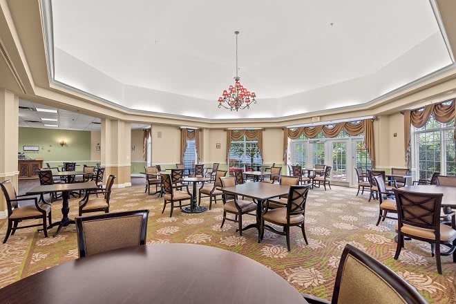 A spacious dining room with elegant furnishings, featuring multiple tables and chairs, a central chandelier, and large windows providing natural light.