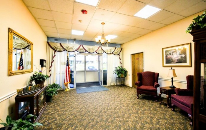 A warmly lit lobby with a carpeted floor, featuring armchairs, potted plants, and an American flag near the entrance.
