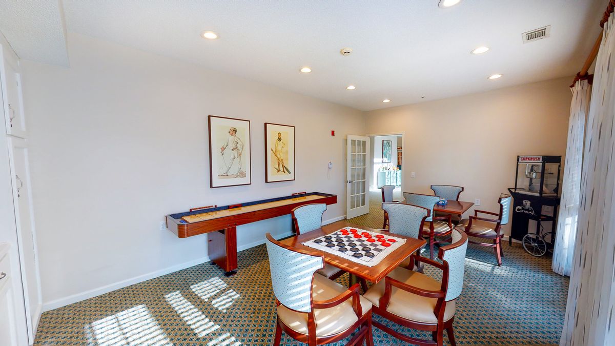A well-lit game room featuring a shuffleboard table and a chessboard with chairs around it.