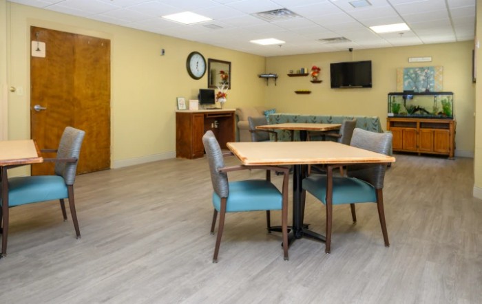 A clean and organized dining or community space with tables, chairs, a television, and a wall clock.
