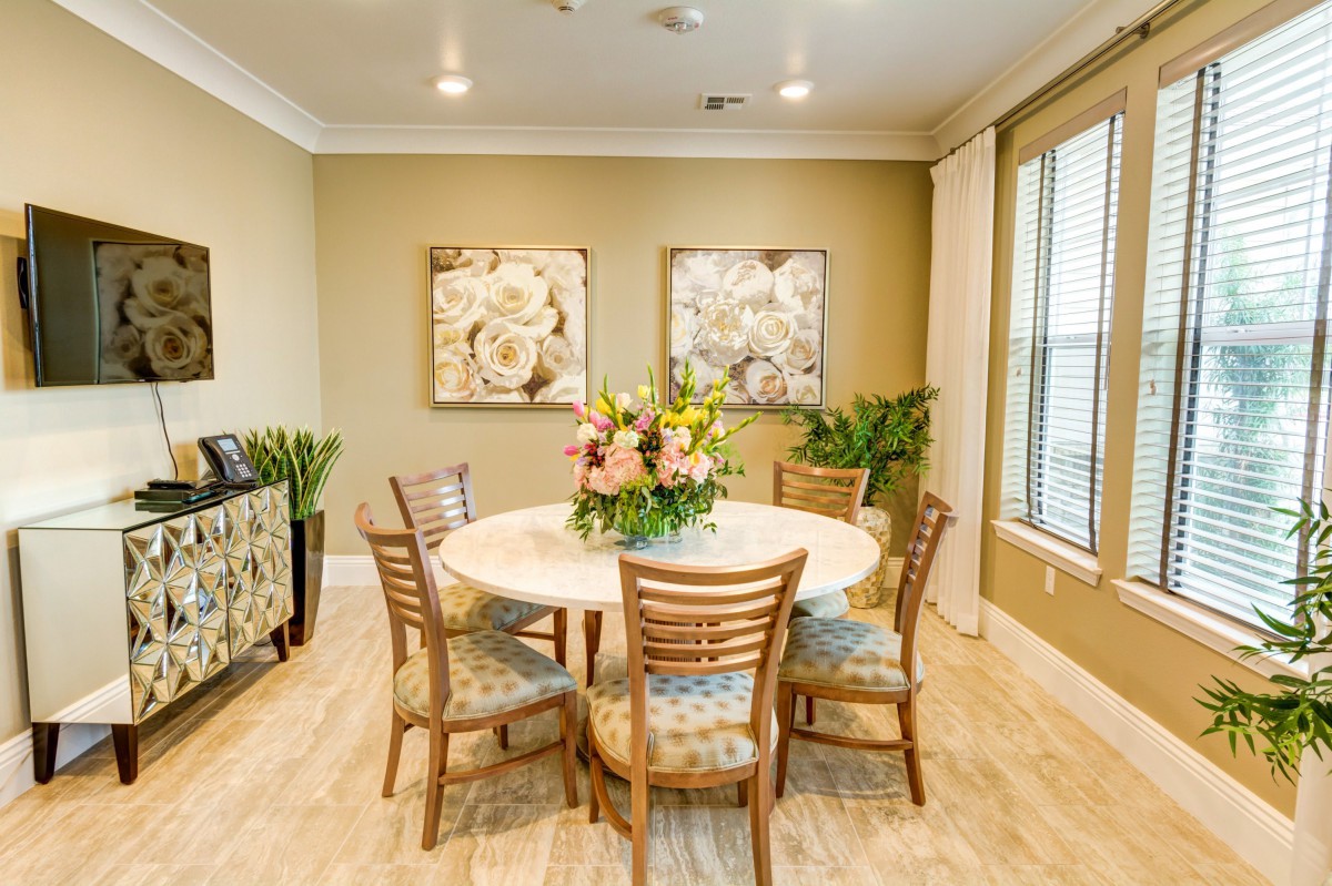 A cozy dining room with a round table surrounded by wooden chairs, featuring a floral centerpiece and decorative art on the walls.