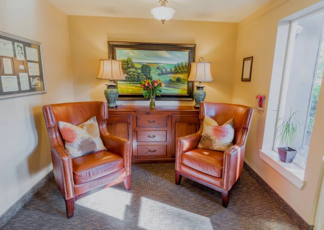 A cozy living room corner featuring two leather chairs, a wooden cabinet, and a painting with lamps on either side.