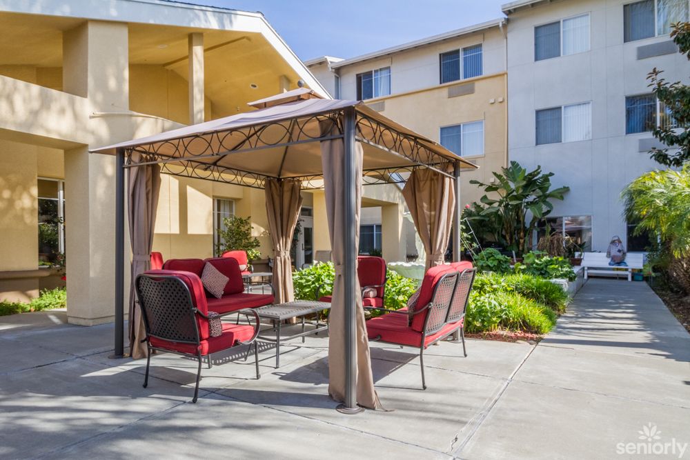 A cozy outdoor seating area with red-cushioned patio furniture under a gazebo in front of an apartment building.