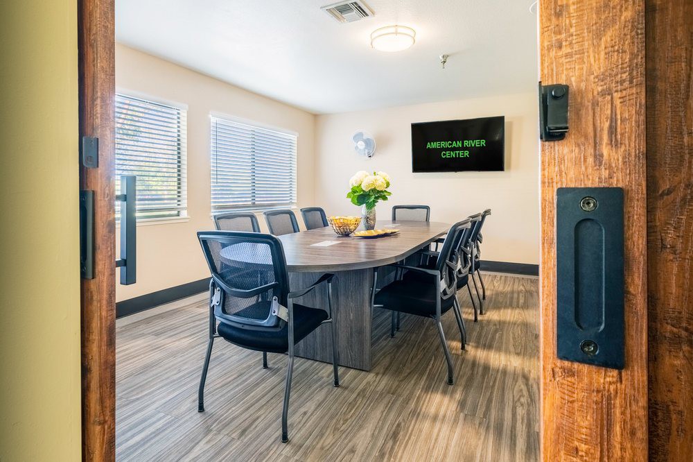A modern conference room with a central table, chairs, and a TV screen, illuminated by natural light.