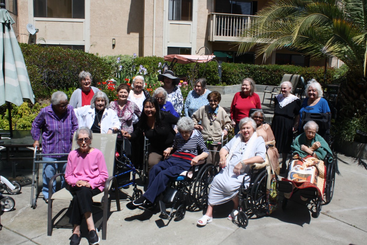 A group of elderly people, some in wheelchairs, enjoying a sunny day outside a retirement home.