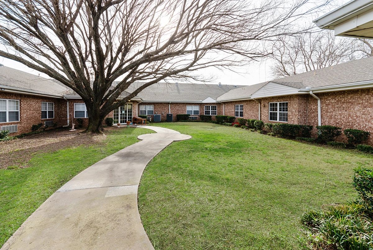 A curved pathway leads through a grassy courtyard with a large tree, surrounded by a brick building.