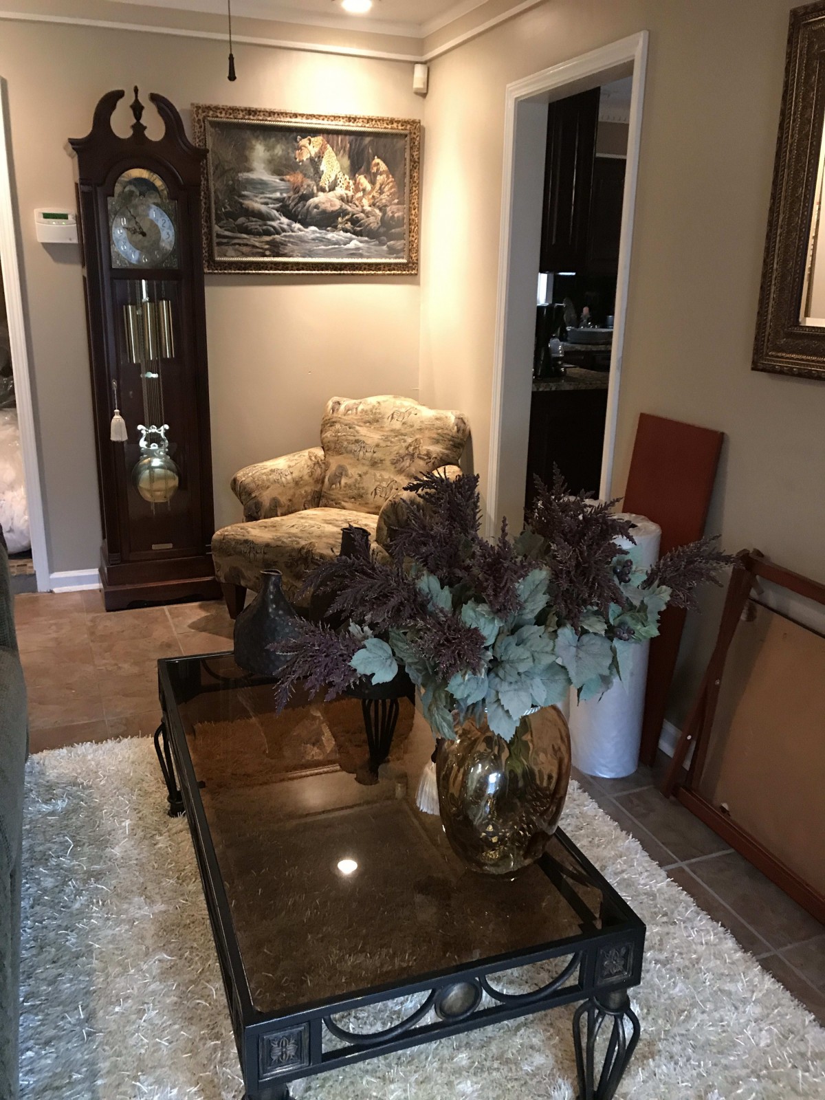 A cozy living room with an armchair, coffee table, and a grandfather clock, decorated with paintings and a vase of flowers.