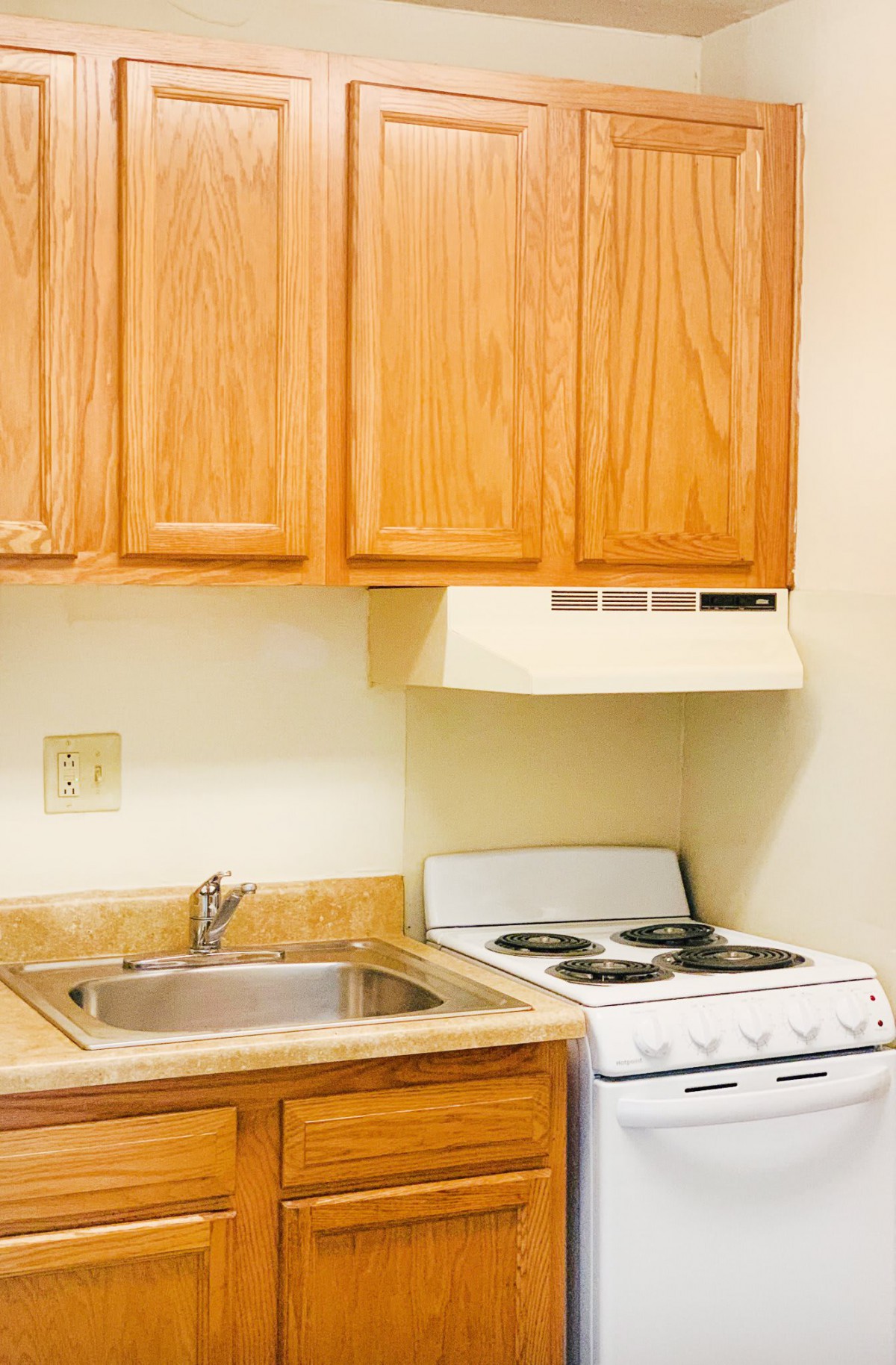A compact kitchen with wooden cabinets, a stainless steel sink, and a stove with an overhead hood.