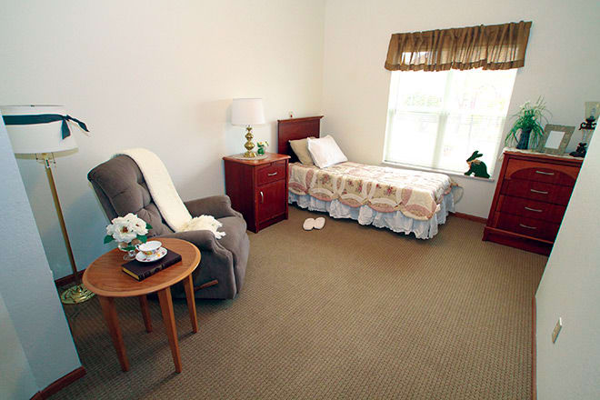 A cozy bedroom featuring a single bed with a bedside table and lamp, an armchair with a side table, and a window with curtains.