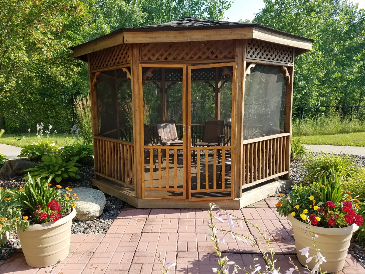 A wooden, screened gazebo sits on a tiled patio amidst lush garden surroundings.