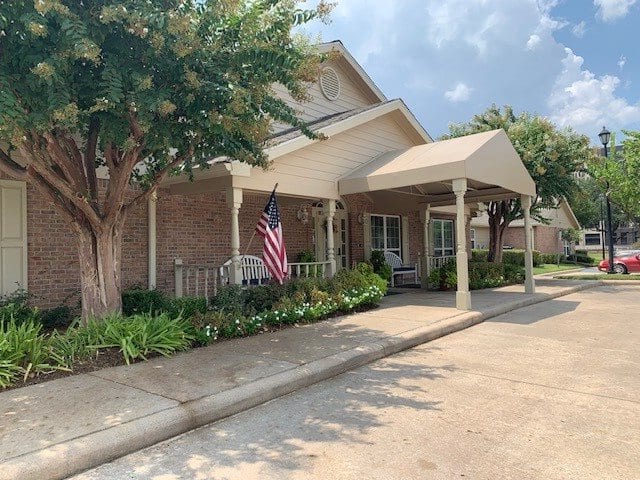 A brick residential house with a covered entrance and an American flag displayed, surrounded by trees and landscaping.