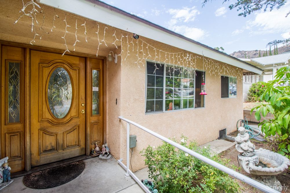 A cozy house exterior with a wooden front door, decorated with string lights above the entrance and a small garden beside the pathway.
