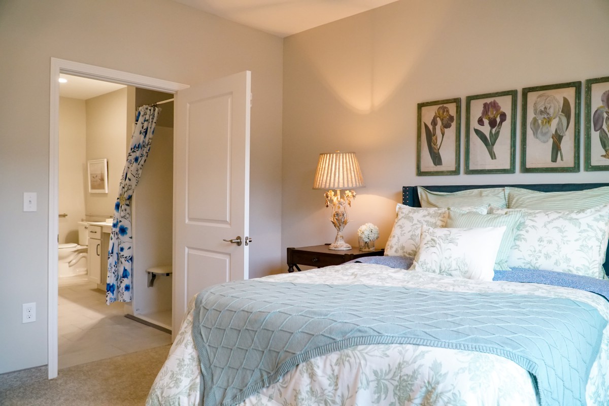 A cozy bedroom with elegant decor, featuring a neatly made bed, stylish lamp, and floral artwork.