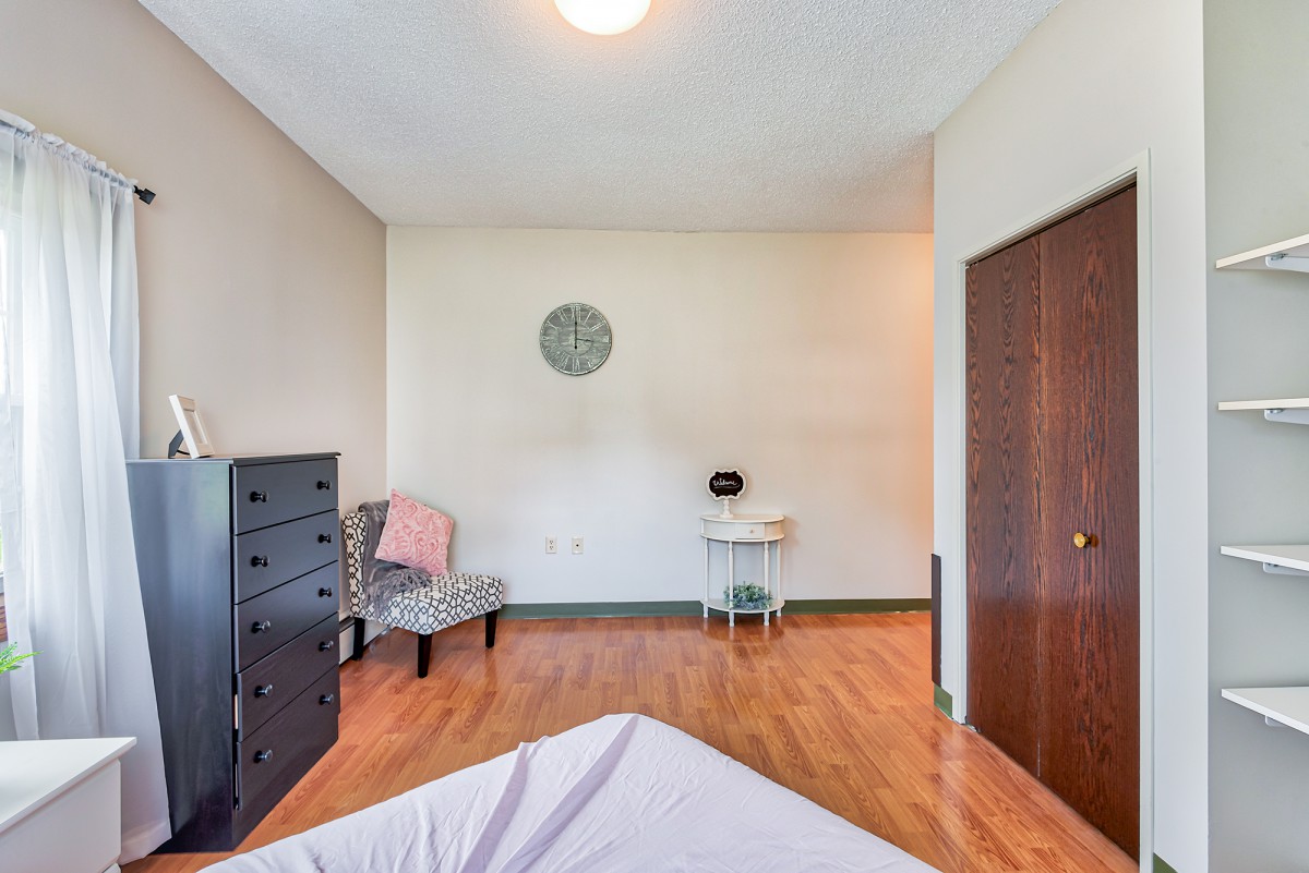 A minimalist bedroom featuring a dresser, chair, and desk with wooden flooring.