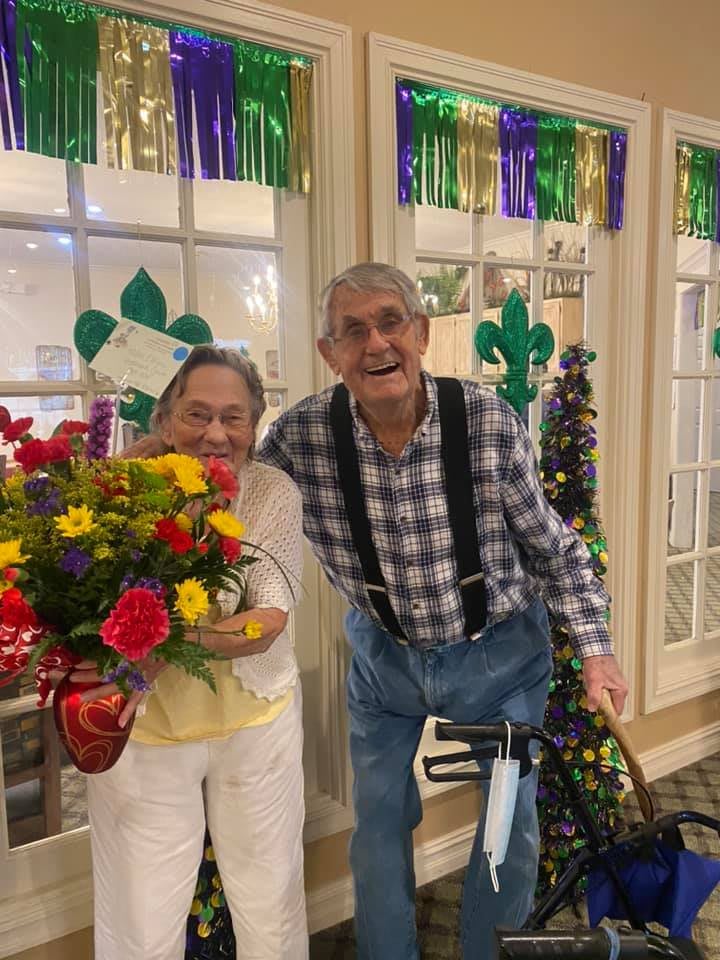 An elderly couple happily posing with a bouquet of flowers, surrounded by festive decorations indoors.
