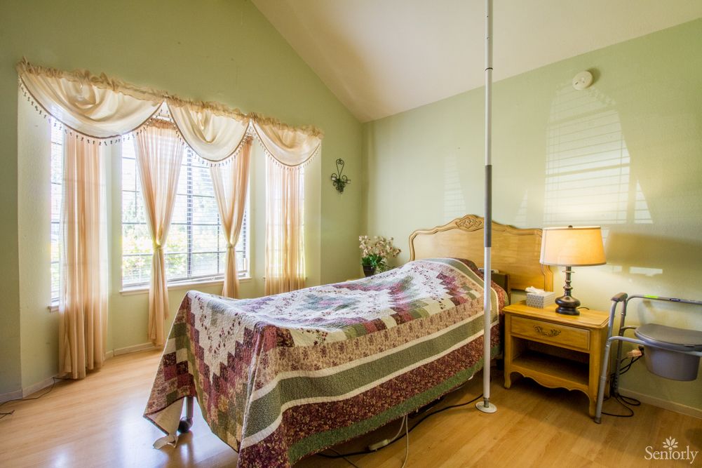A cozy bedroom with a quilt-covered bed, large windows with curtains, and a bedside lamp on a wooden floor.