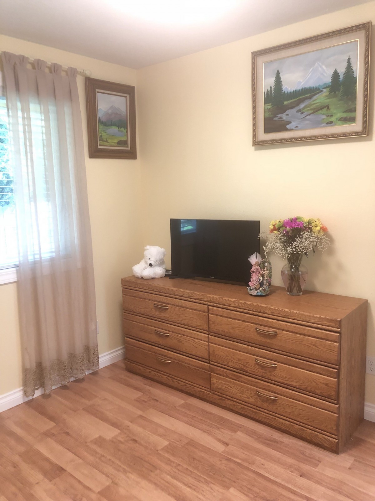 A cozy bedroom corner featuring a wooden dresser topped with a television, decorative items, and flowers, alongside a window with sheer curtains and framed paintings on the wall.